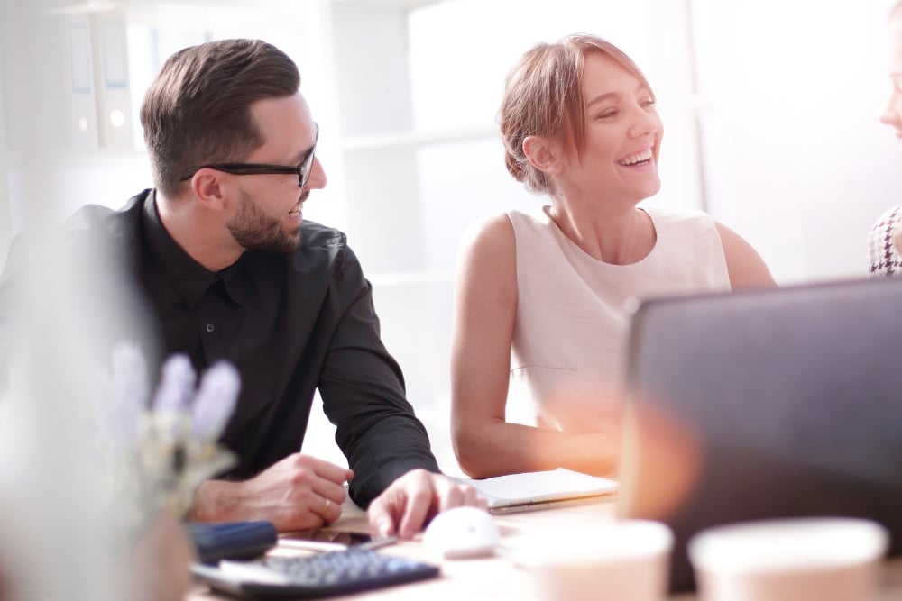 Two employees smiling at a coworker sitting at a desk in the office