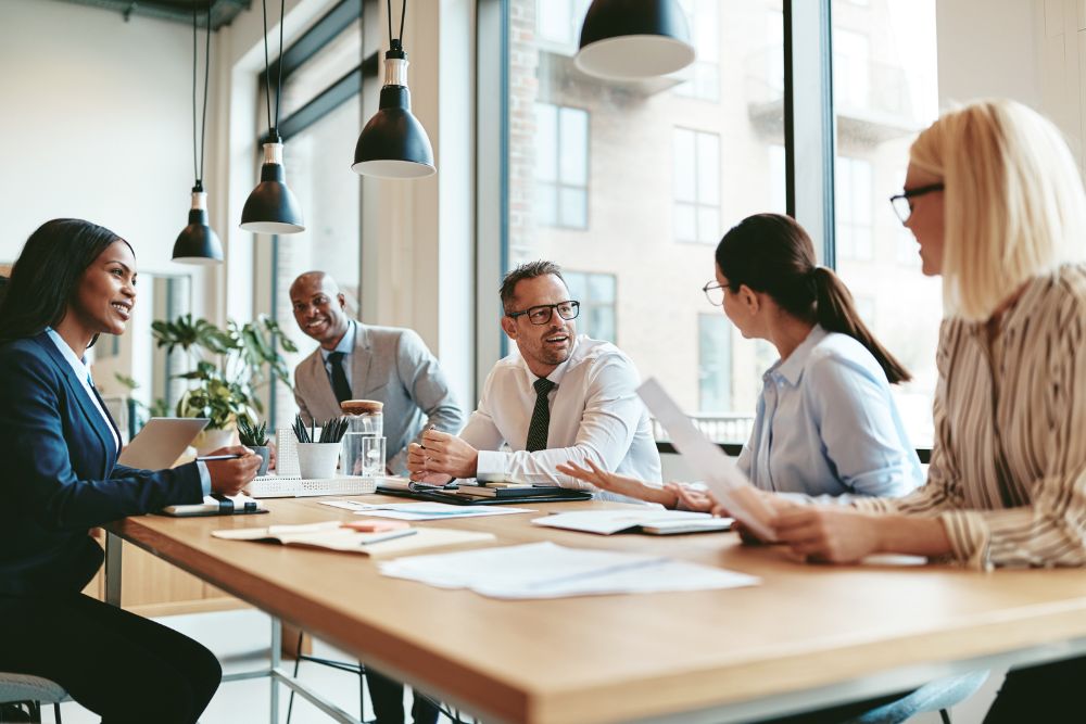 five business people at a desk in the office talking and smiling