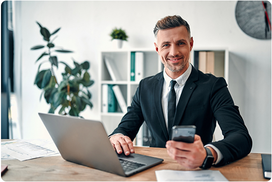 Business man smiling sitting at a desk with a laptop and a phone in one hand