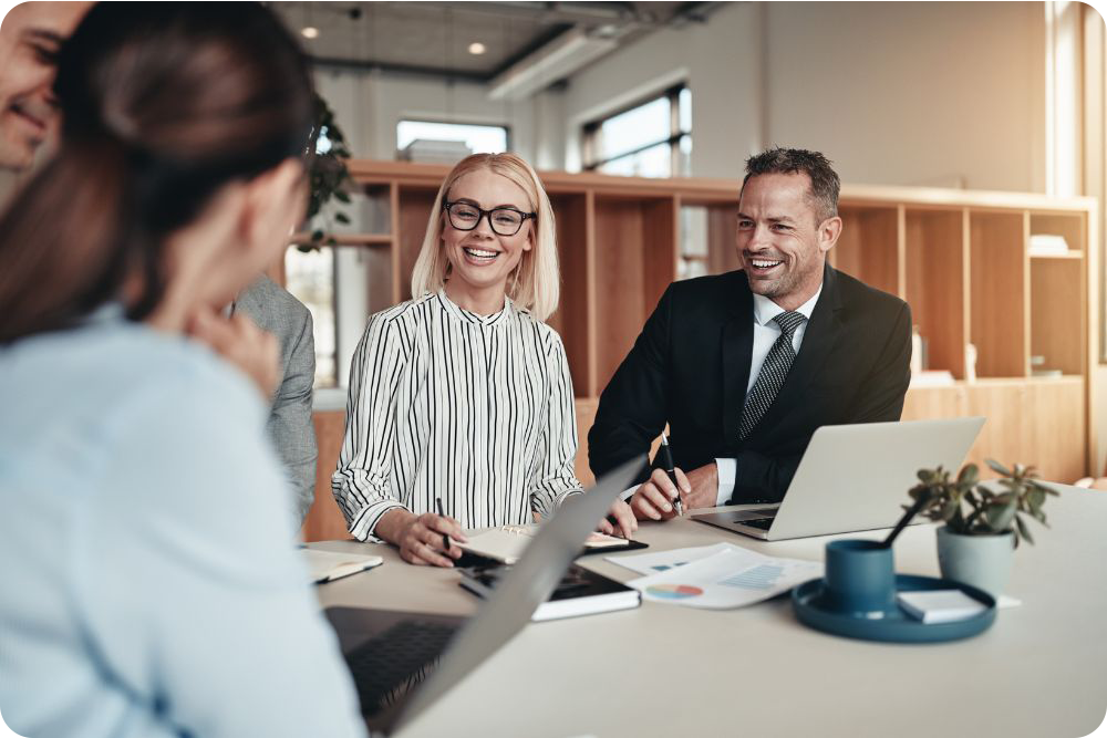 five co-workers smiling with two laptops, notes and a plant at a desk in an office